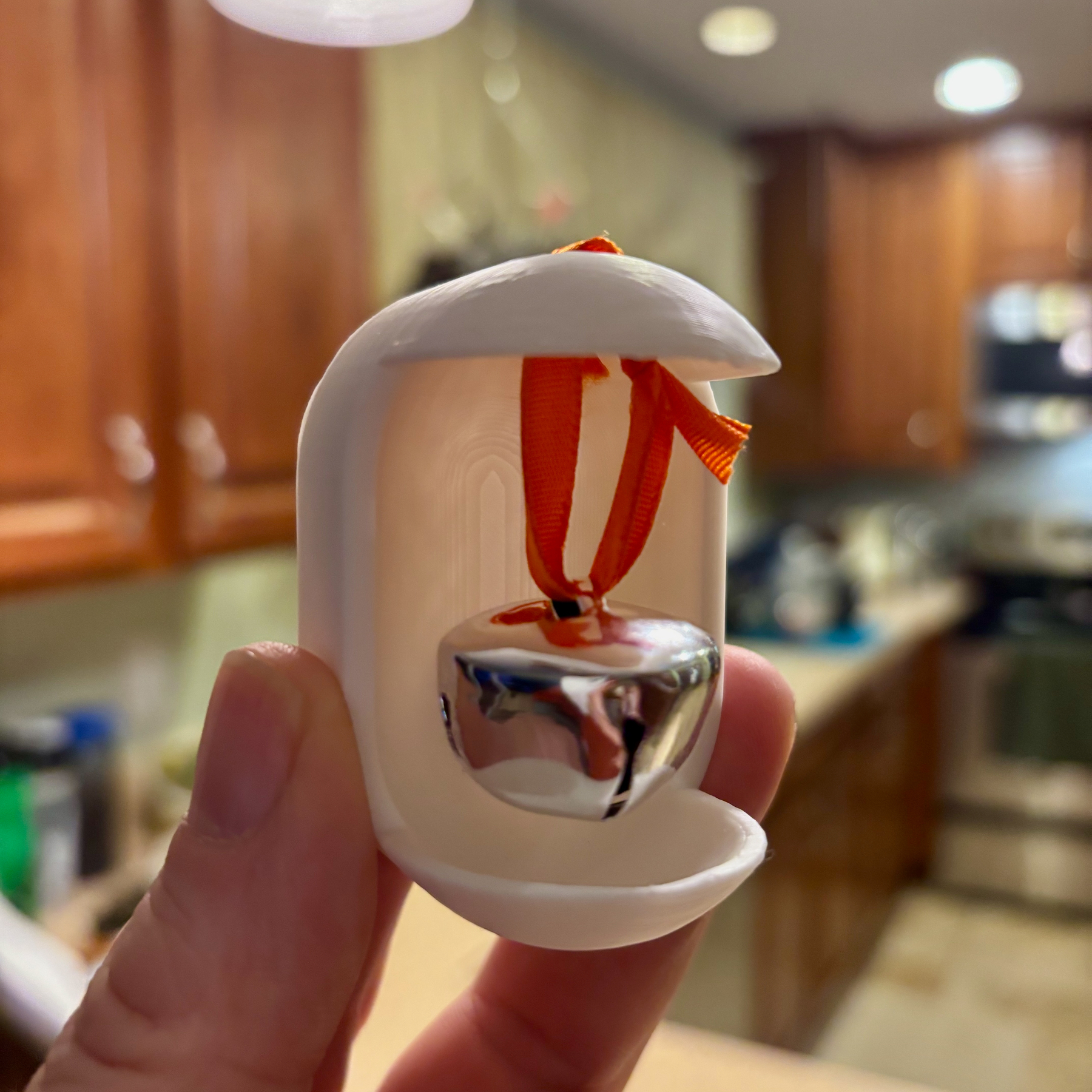 A small silver bell is suspended inside a white plastic holder with an orange ribbon, held by a person's hand in a kitchen setting.