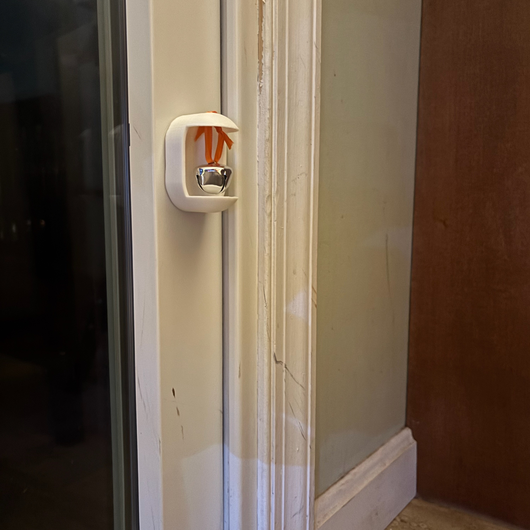 A small silver bell is suspended inside a white plastic holder with an orange ribbon, mounted to a sliding glass door.